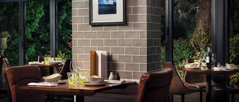 A dining area with brick walls, framed artwork, and a view of greenery through large windows.