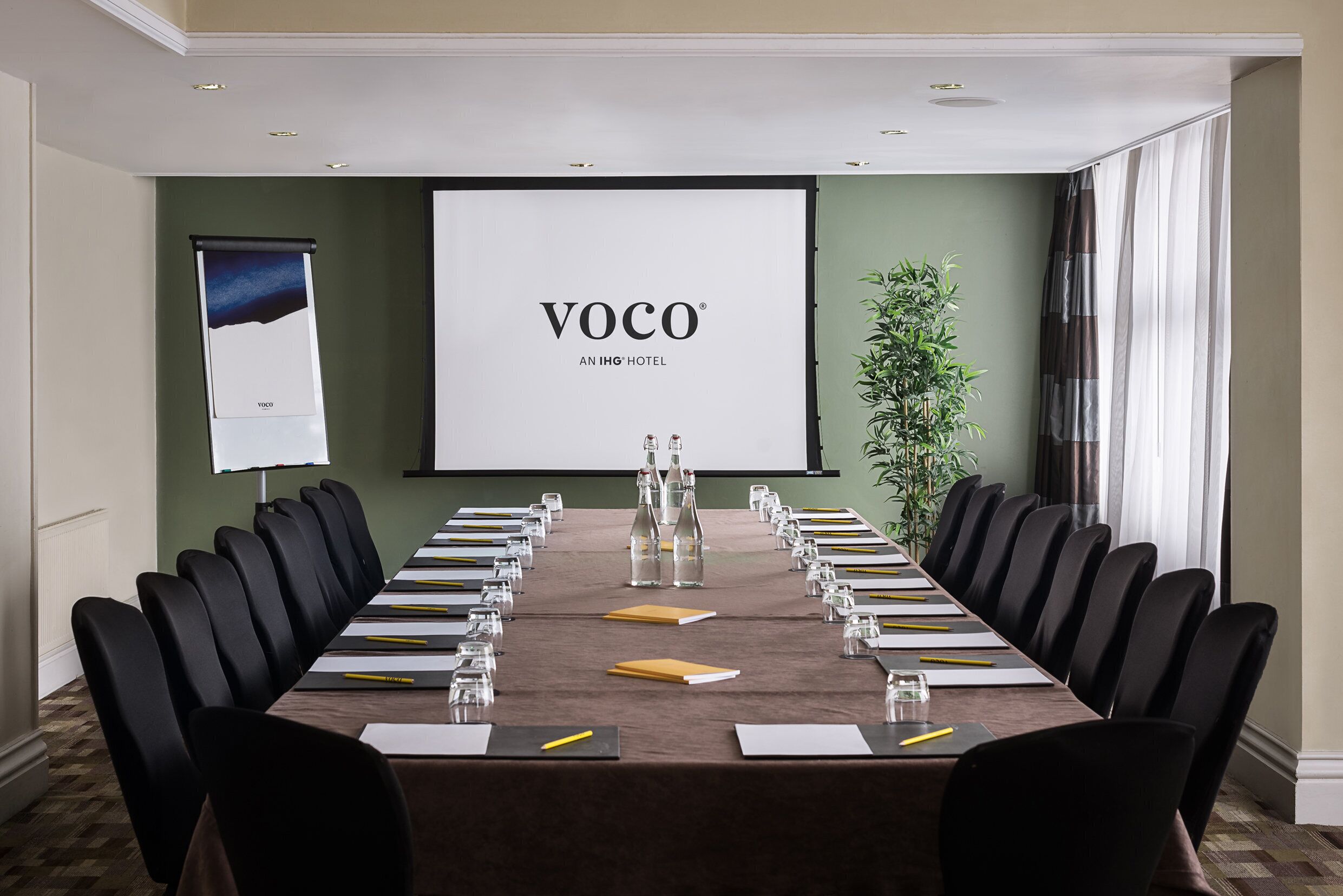 A conference room with a long table, chairs, a projector screen, and a plant.