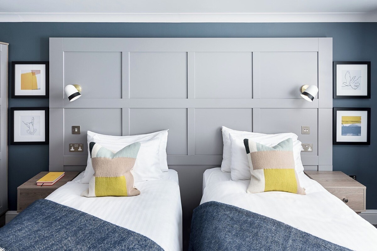 A hotel room with two beds, a grey headboard, bedside lamps, and framed artwork on the walls.