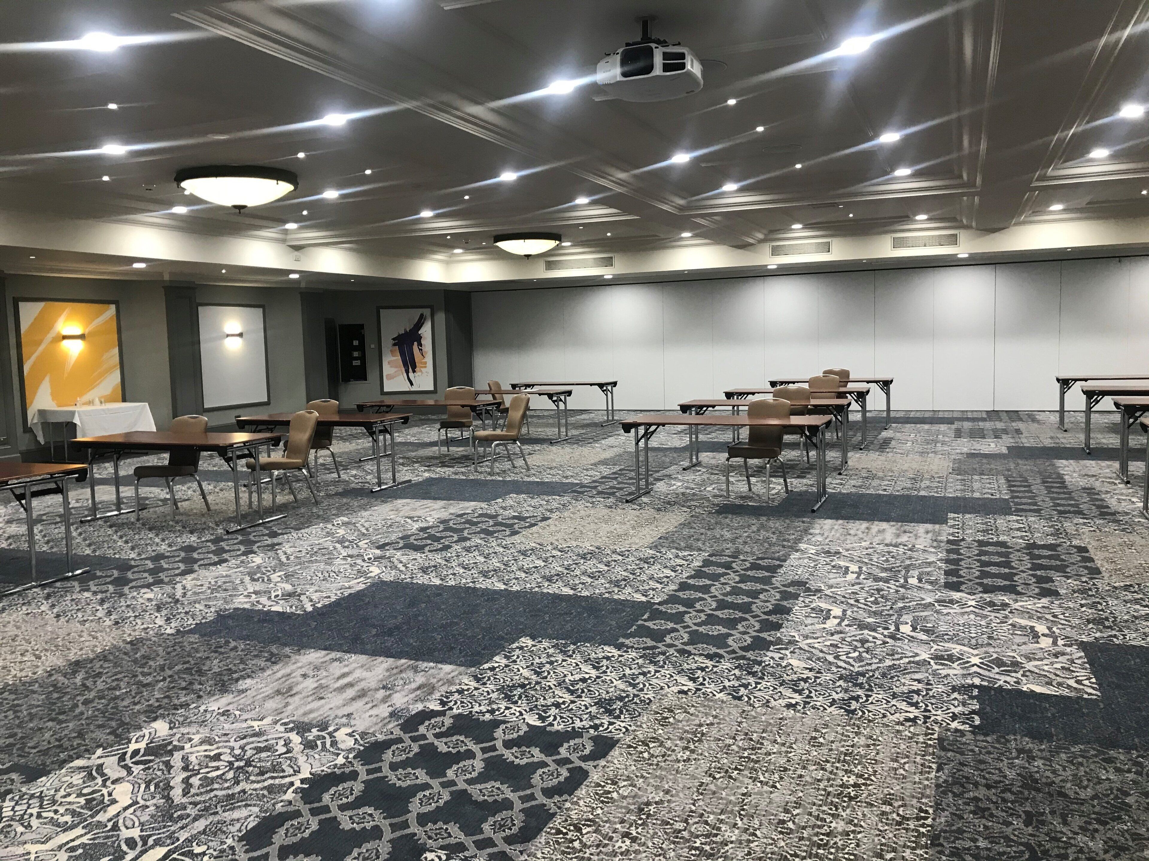 Une salle de conférence équipée de tables et de chaises, dotée d’un sol recouvert de moquette et d’un projecteur fixé au plafond.