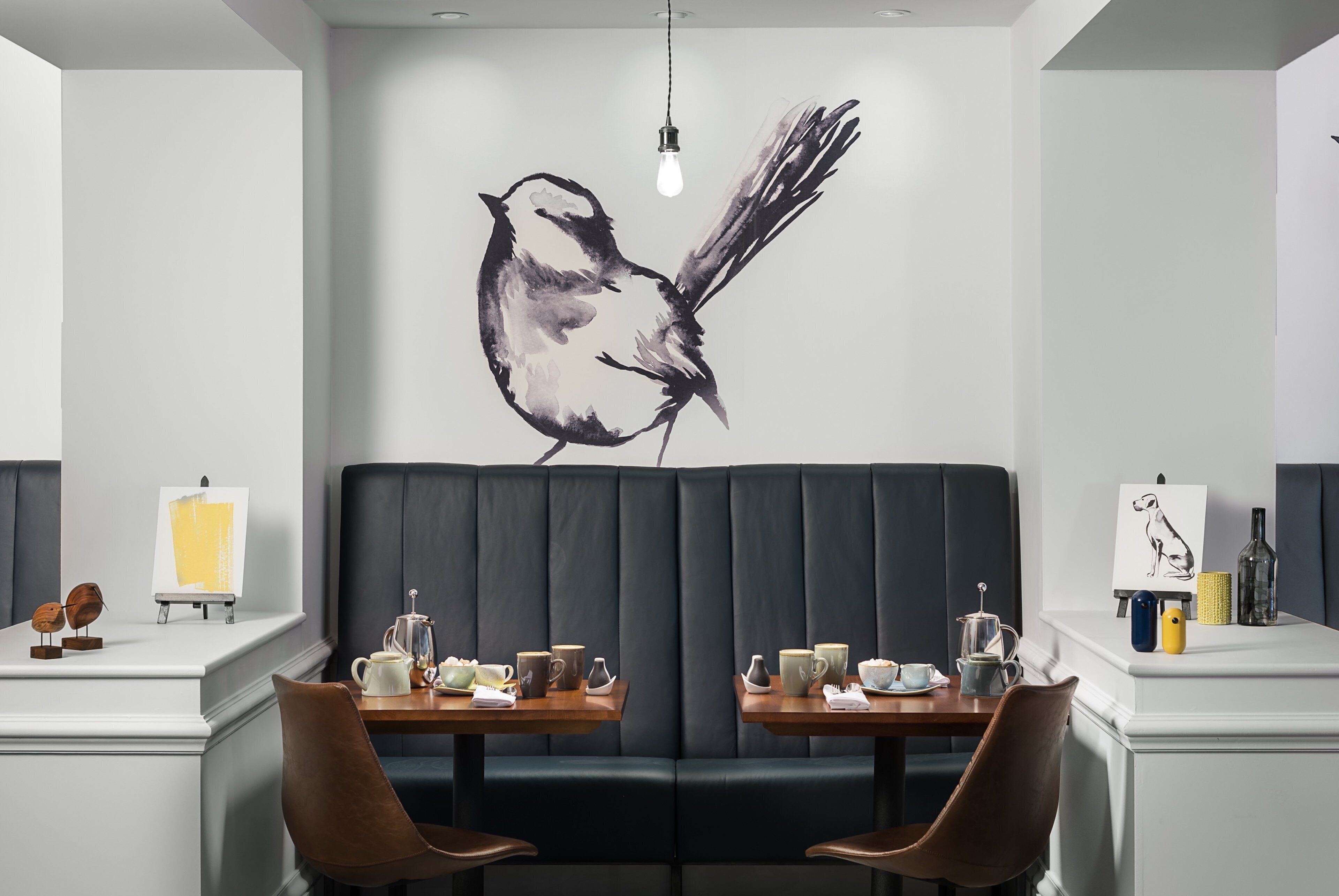 Un comedor moderno con un gran mural de un pájaro, dos mesas con sillas y una lámpara colgante.