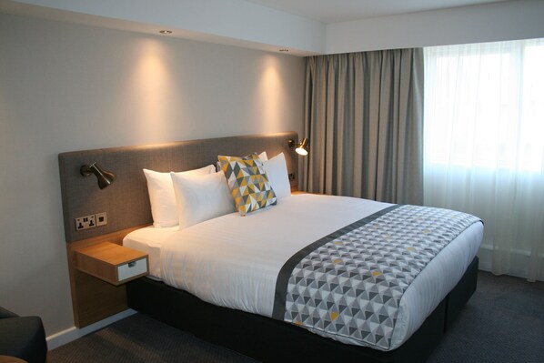Una habitación de hotel con una cama grande, una mesita de noche, una lámpara y un ventanal con cortinas.