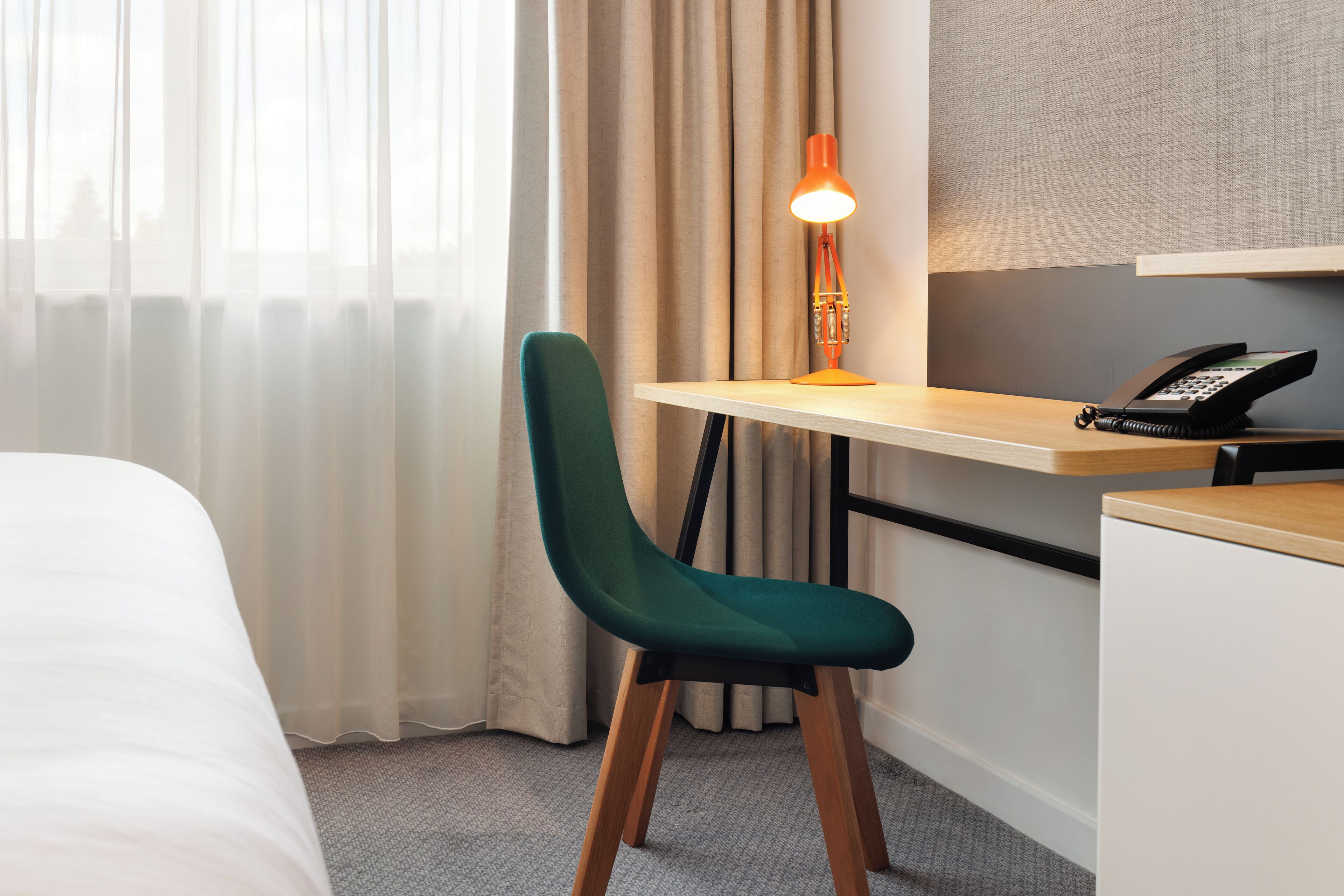 A hotel room with a bed, a desk with a telephone, a chair, and a lamp.