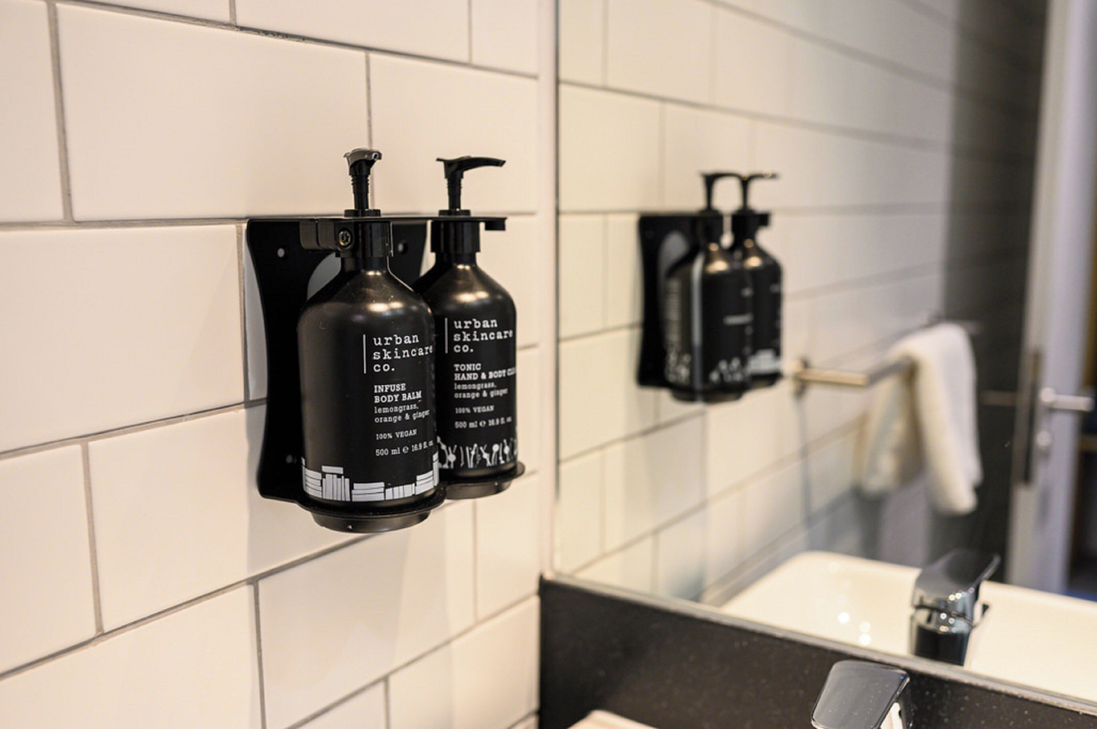 Casa de banho com parede revestida a azulejos brancos, bancada preta e dois dispensadores pretos com a inscrição 'urban skincare co'.