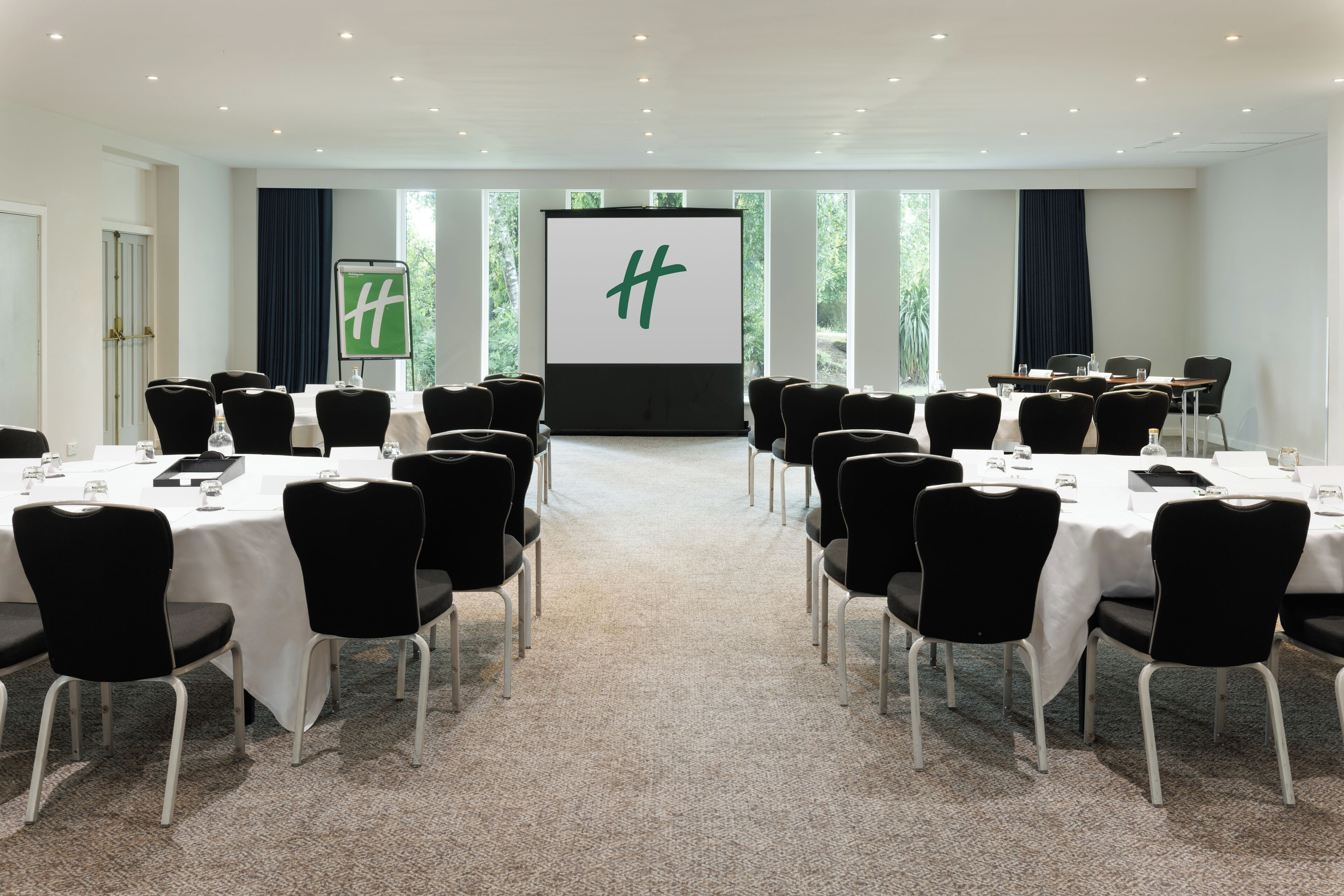 Une salle de conférence dotée d’un grand écran affichant un logo, de chaises noires, de tables blanches et d’un sol recouvert de moquette.