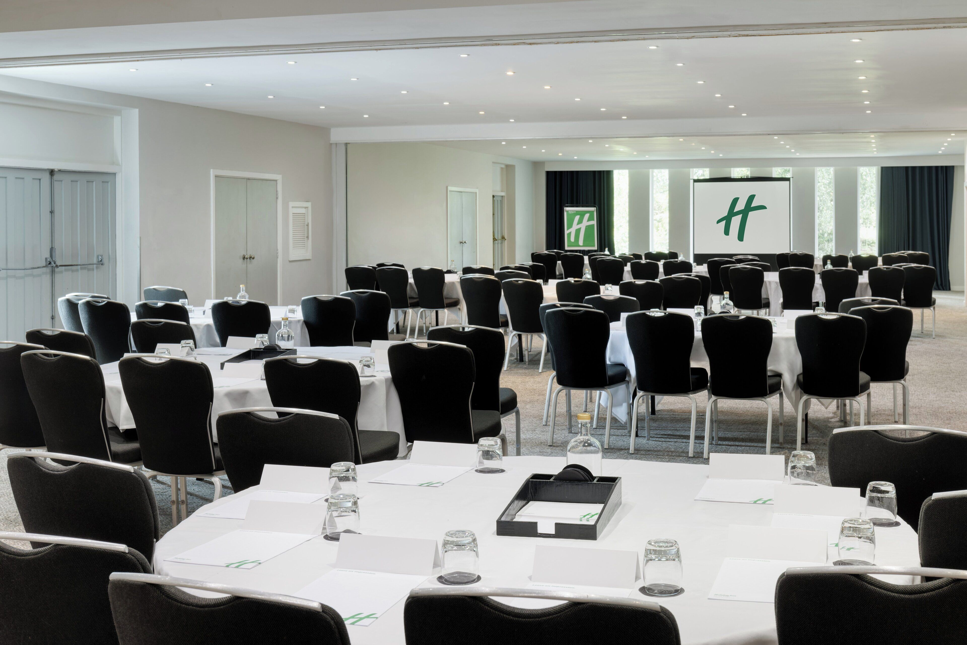 A conference room with multiple tables and chairs, a large screen, and a door with a green logo.