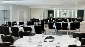 A conference room with multiple tables and chairs, a large screen, and a door with a green logo.