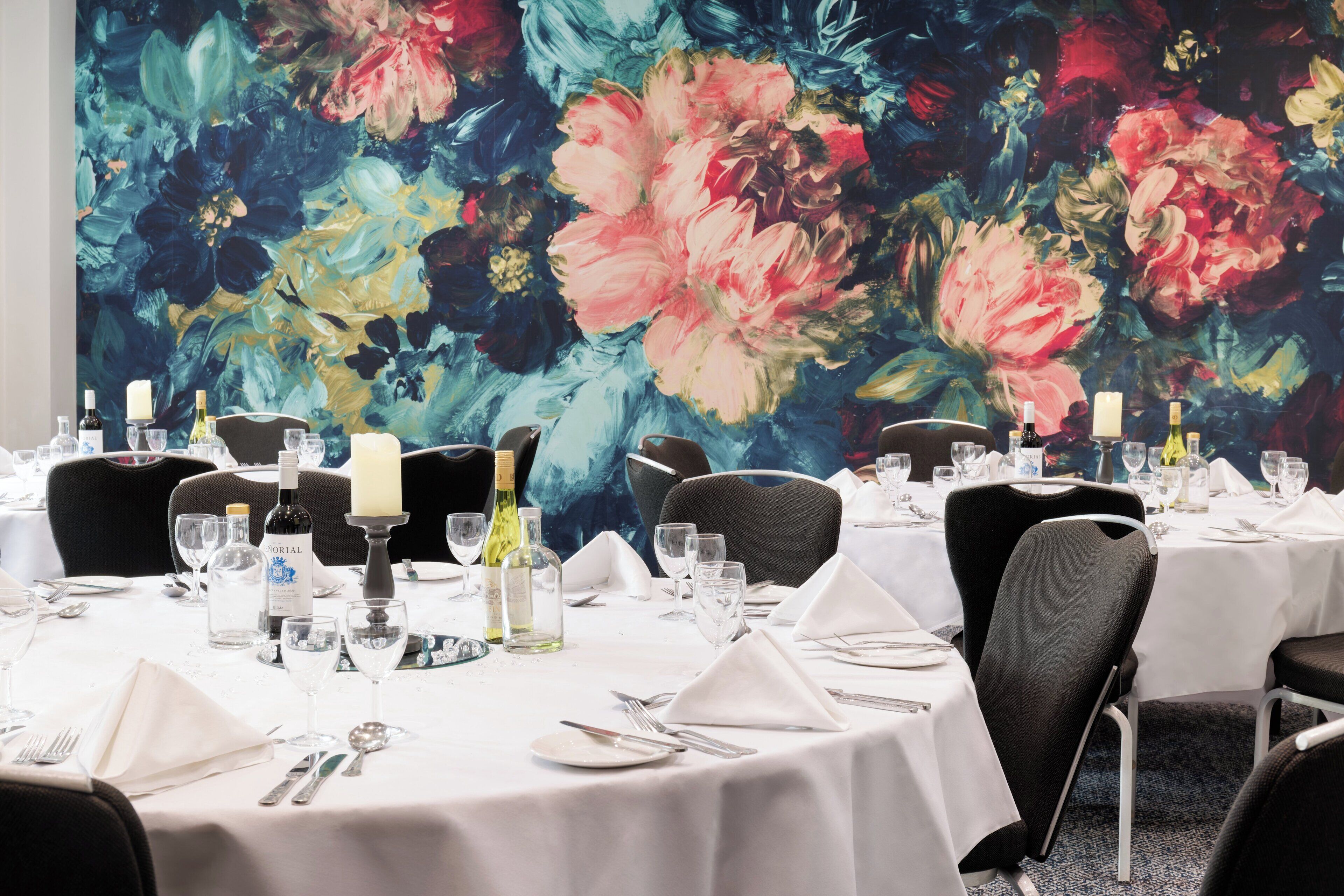 Un comedor con mesas redondas dispuestas para una comida, con manteles blancos, sillas negras y un cuadro grande con motivos florales en la pared.