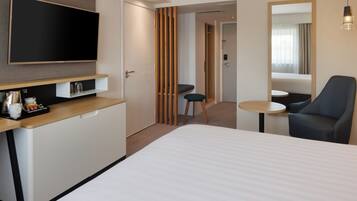 A modern hotel room with a bed, a desk with a flat-screen TV, a small round table, and a chair.