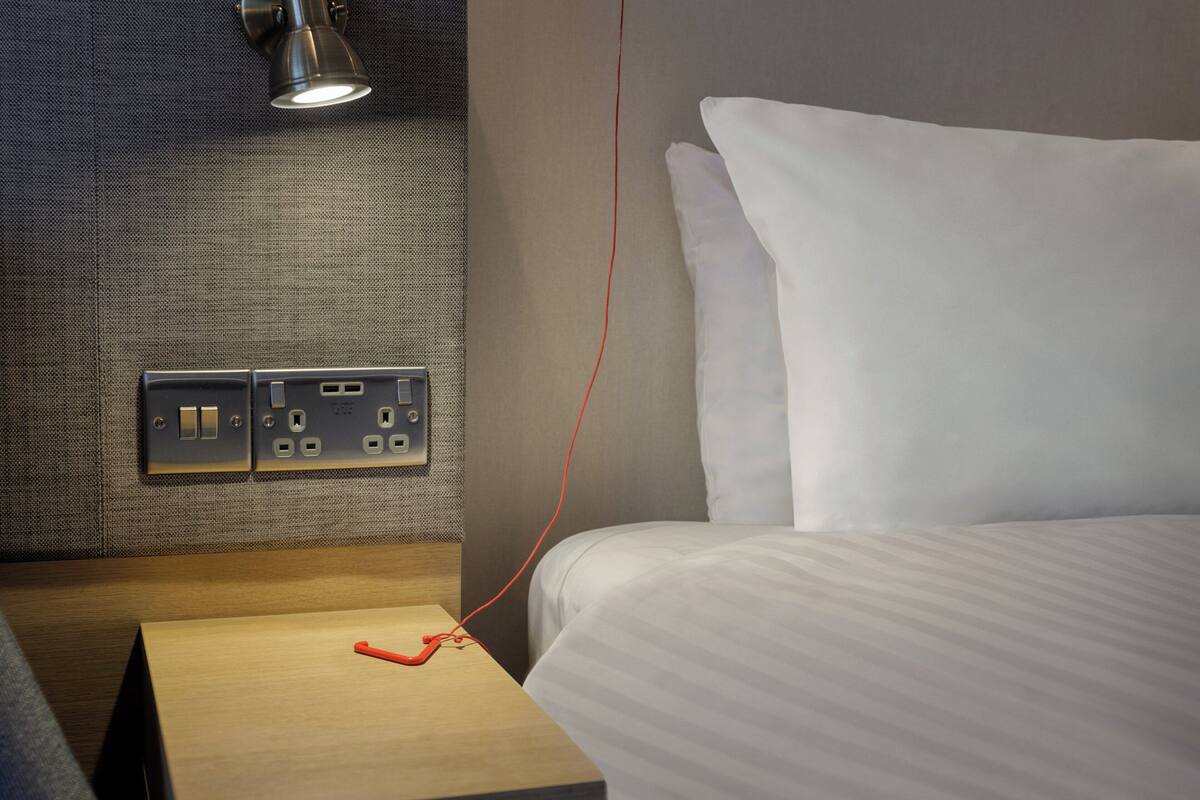 A hotel room with a bed, a nightstand, and a wall socket with a red cable plugged in.