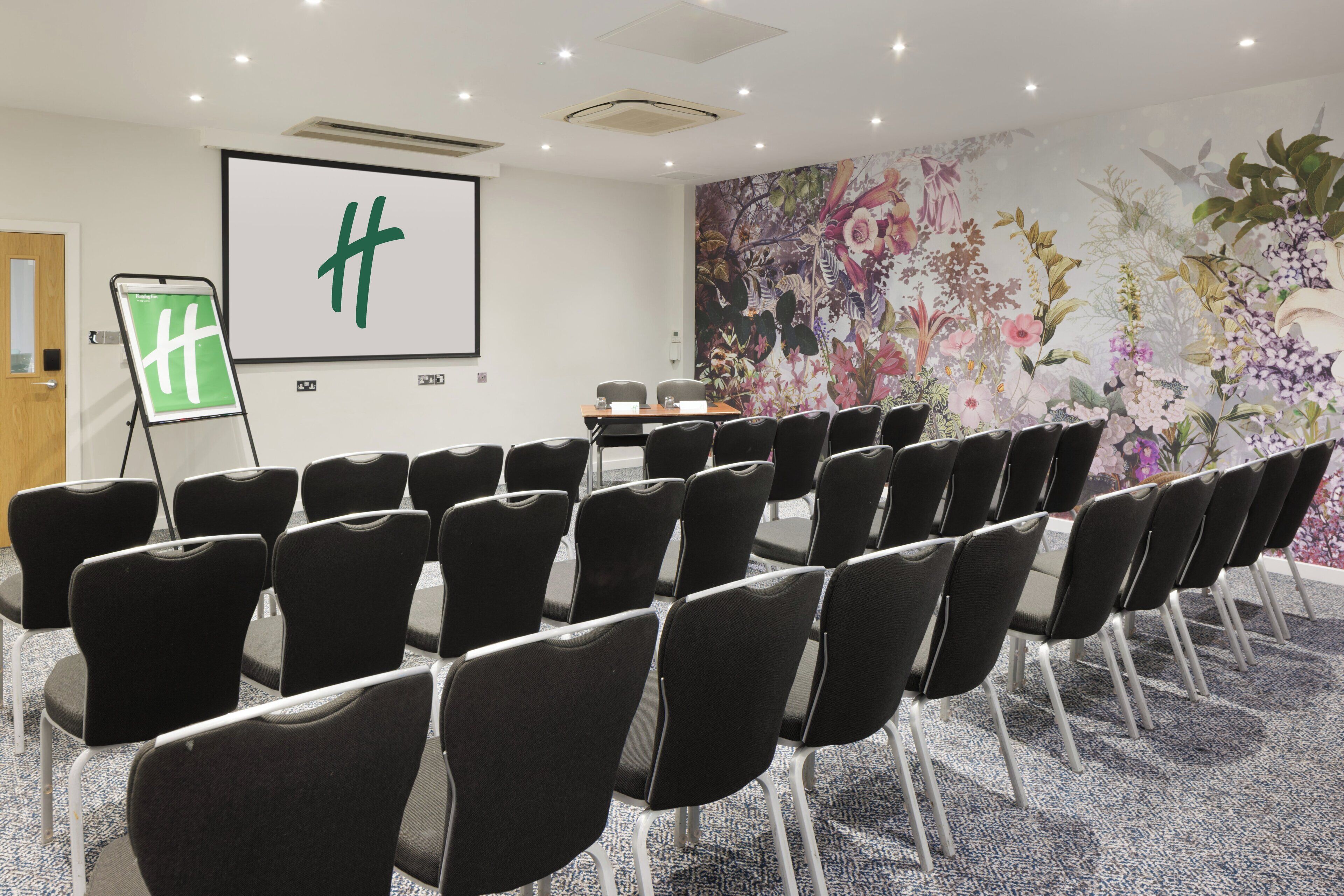 Une salle de conférence avec des rangées de chaises noires, un écran de projection orné d’un logo « H » vert, une fresque florale et un sol recouvert de moquette.