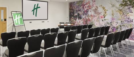 Una sala de conferencias con filas de sillas negras, una pantalla de proyector con el logotipo de una 'H' verde, un mural floral y piso alfombrado.
