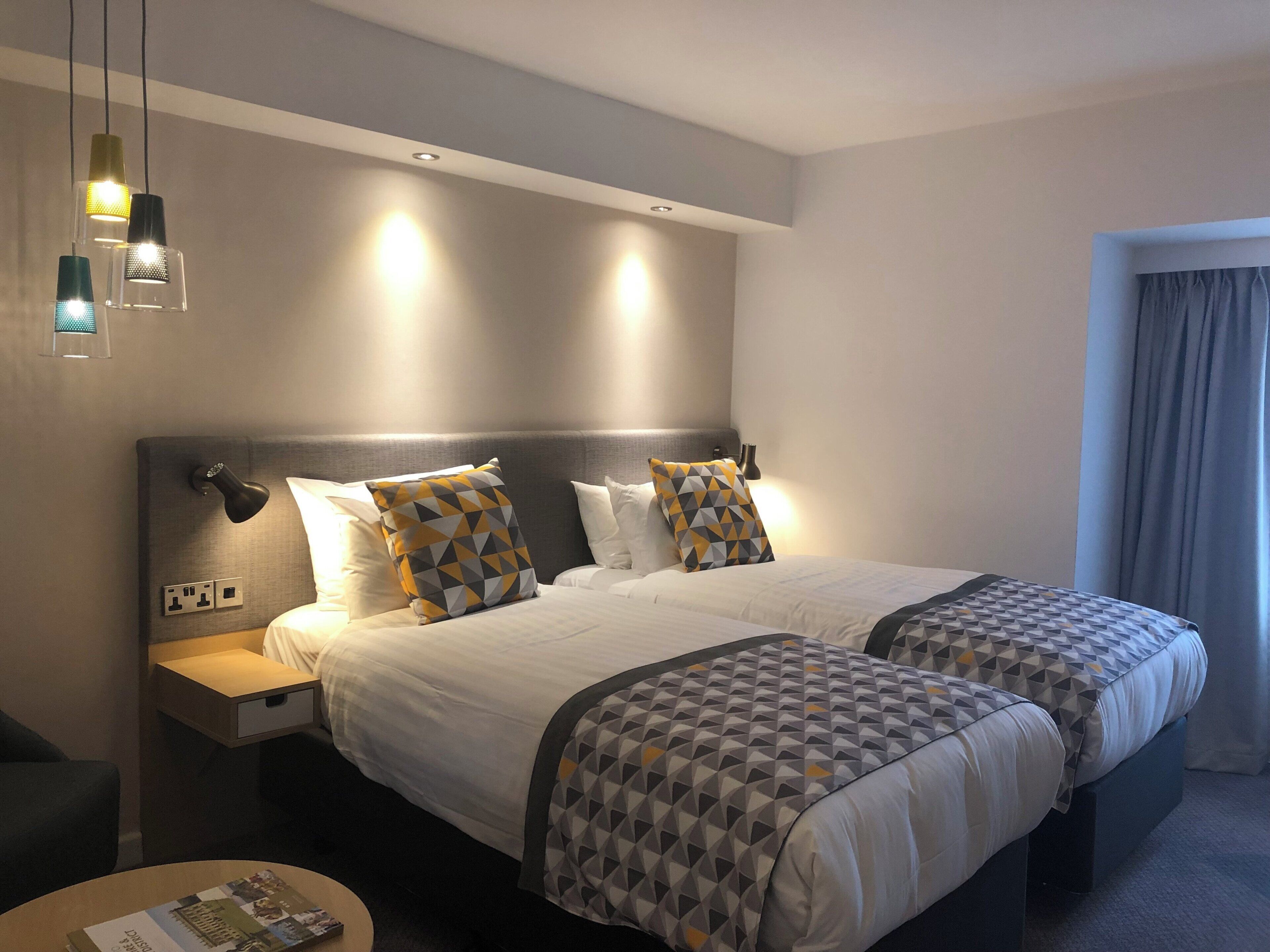 Habitació d'hotel amb dos llits, una taula de nit, una làmpada i una finestra amb cortines.