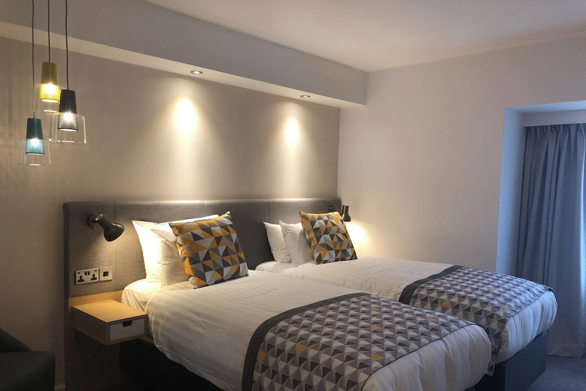 A hotel room with two beds, a bedside table, a lamp, and a window with curtains.