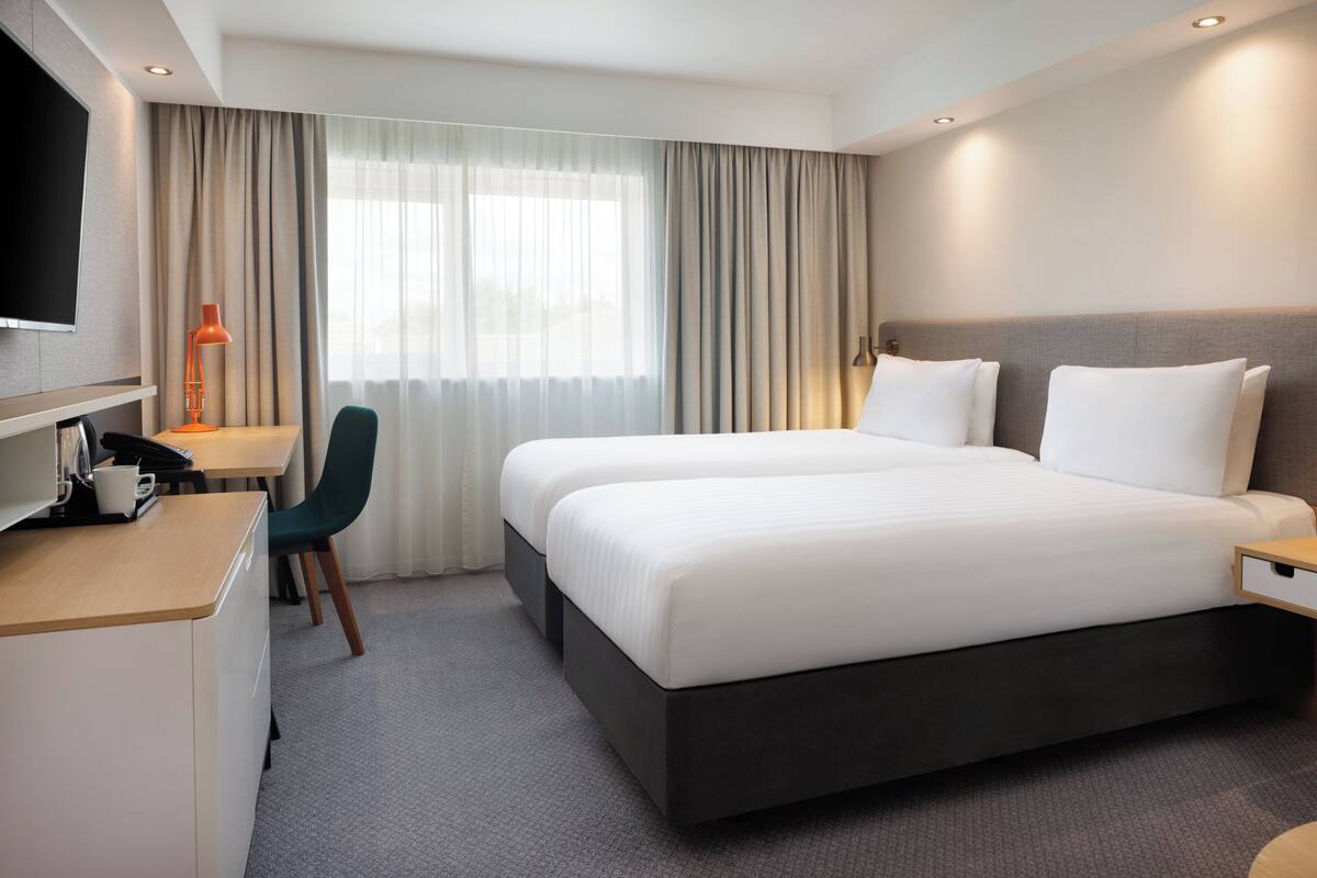 A modern hotel room with a large bed, a desk with a chair, a flat-screen TV, and a large window with curtains.