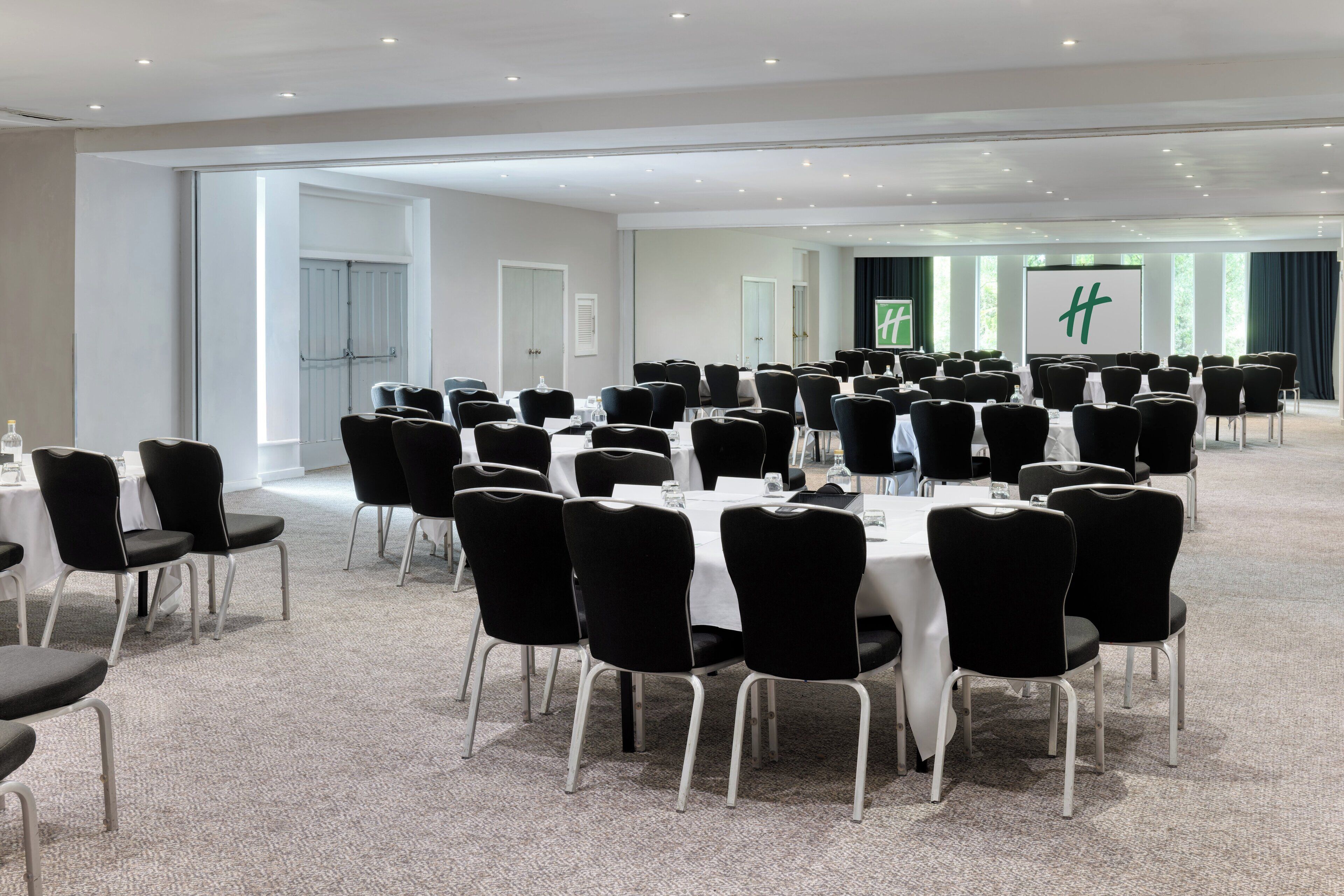 A conference room with round tables and black chairs, large windows, and a stage with a green logo.