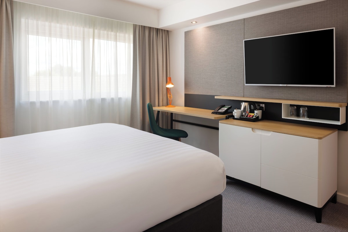 A hotel room with a bed, a desk with a telephone, a flat-screen TV, and a window with curtains.