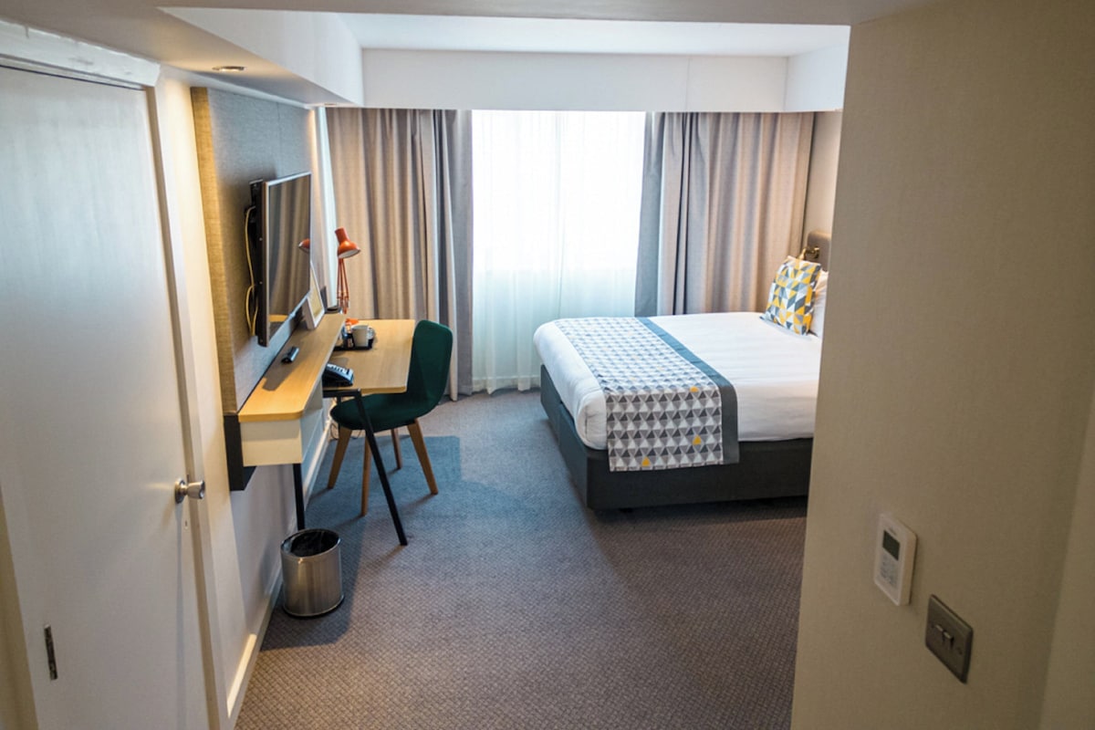 A hotel room with a bed, a desk with a chair, a television, and a window with curtains.