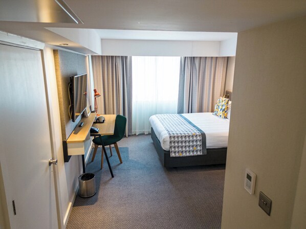 A hotel room with a bed, a desk with a chair, a television, and a window with curtains.