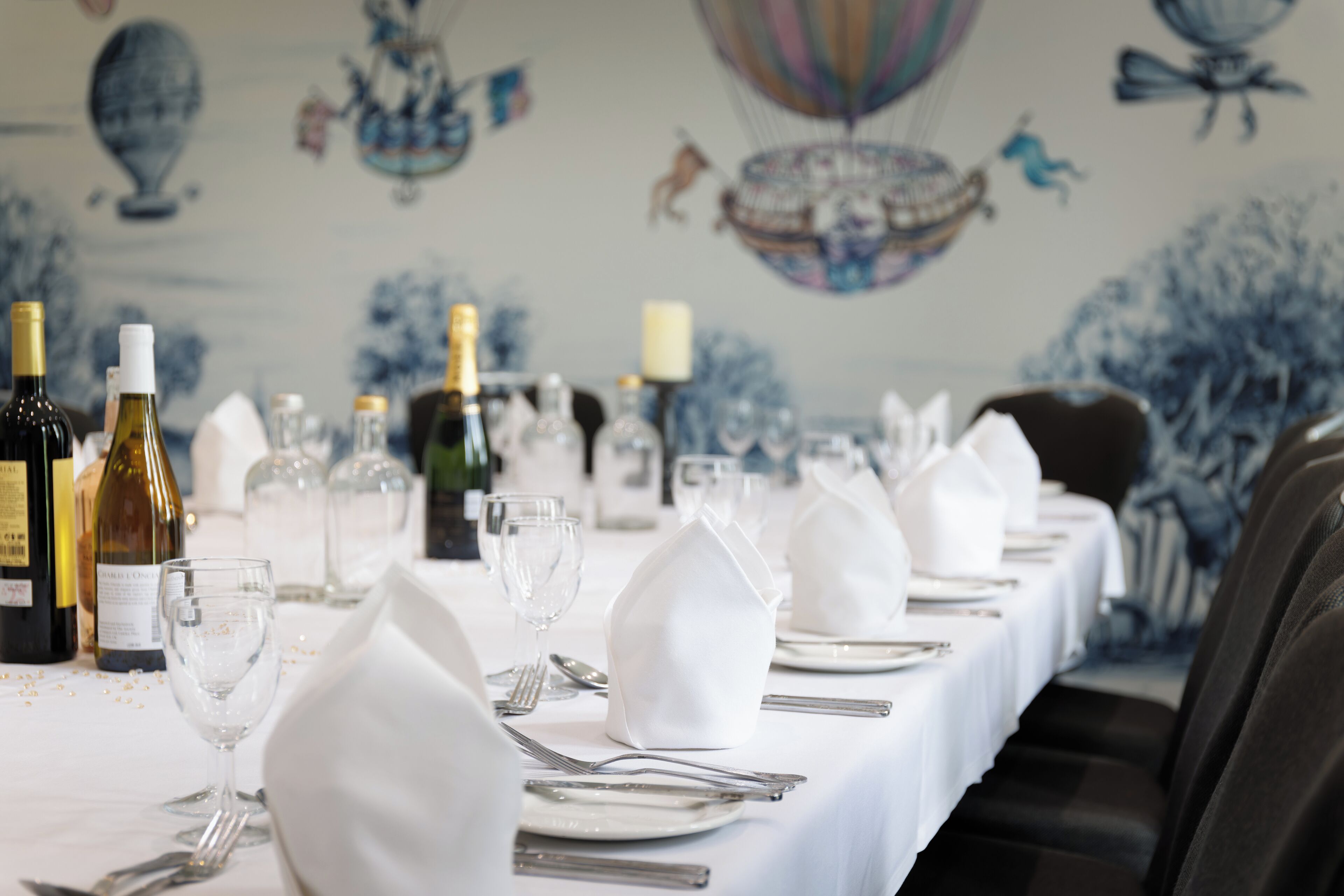 Una mesa de comedor dispuesta con manteles blancos, cubertería y botellas de vino, con un mural de fondo.