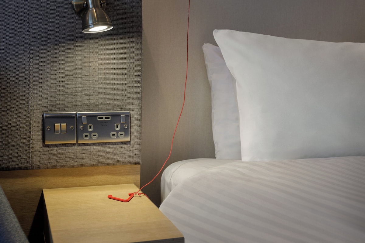 A hotel room with a bed, a nightstand, a lamp, and a power outlet.