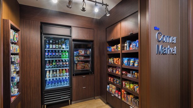 Una tienda de barrio con un mostrador refrigerado para bebidas, estantes con aperitivos y un letrero que dice 'The Corner Market'.