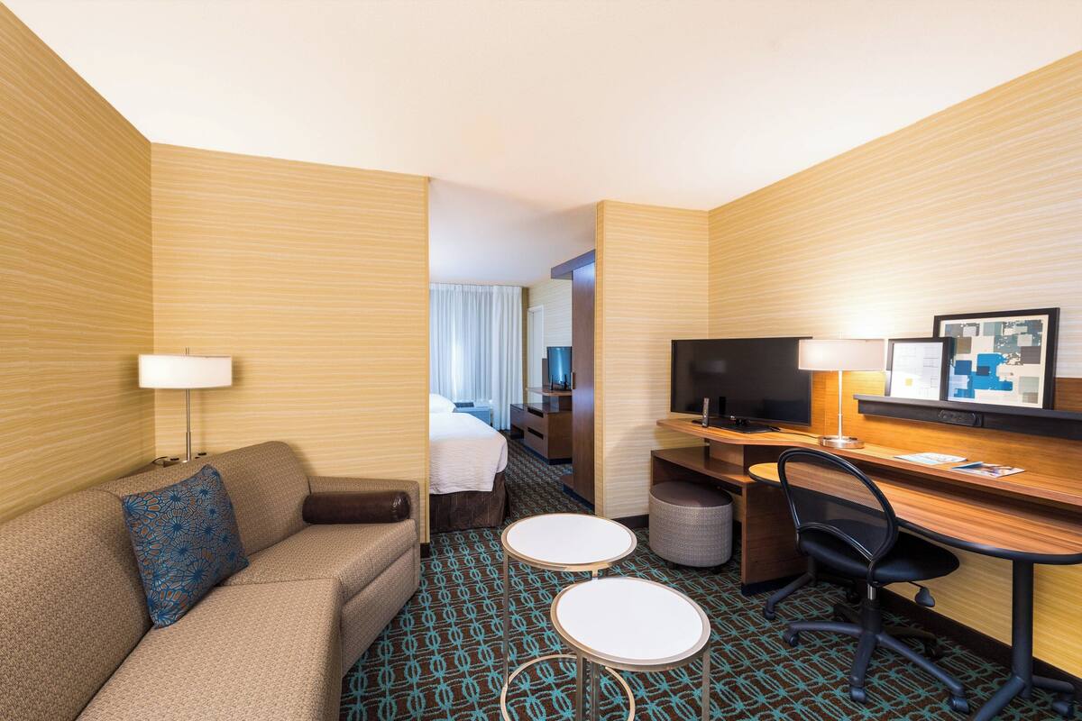 A hotel room with a sofa, two small round tables, a desk with a chair, a TV, and a bed with curtains.