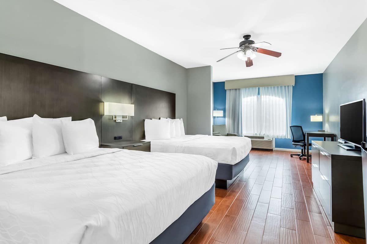 A hotel room with two beds, a desk, a chair, a TV, and a ceiling fan.