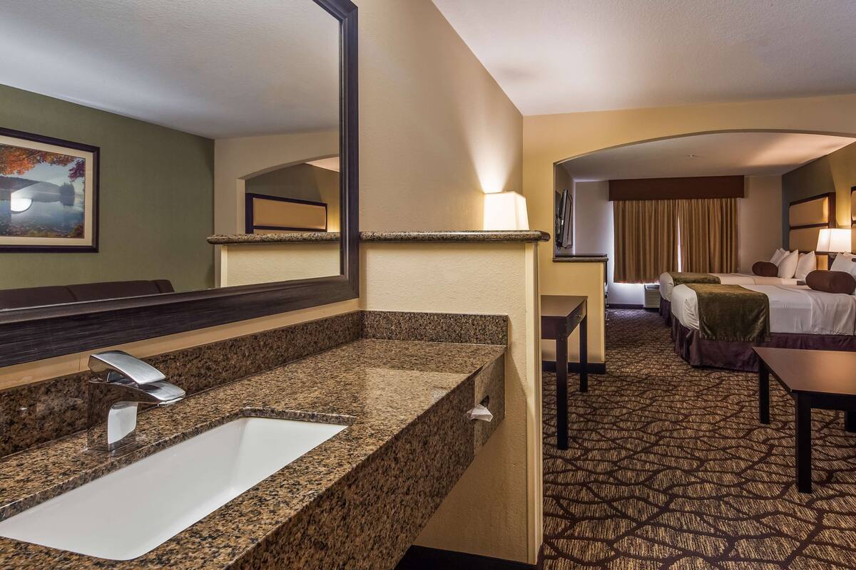 Habitación de hotel con lavamanos de encimera de granito, un espejo grande, una cama con cabecero, una mesita pequeña y un sofá.