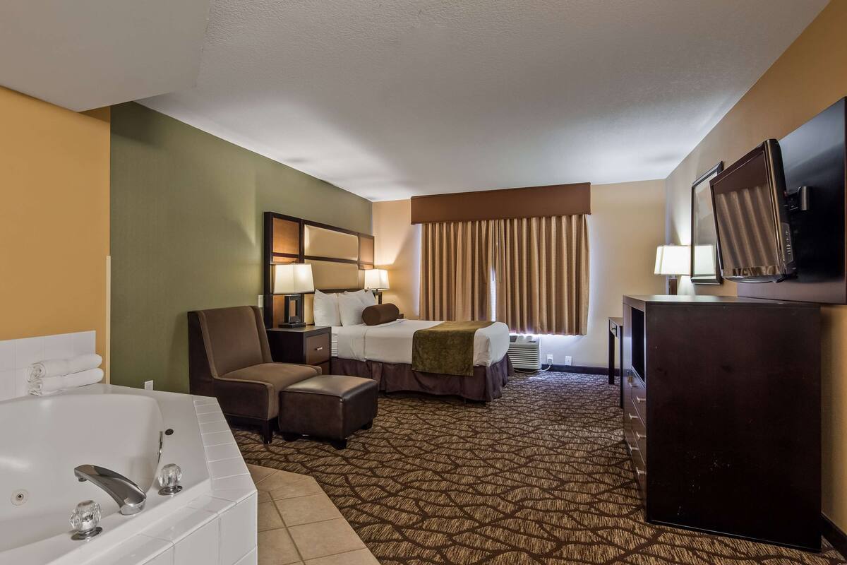 Habitación de hotel con una bañera grande, cama con dos almohadas, una silla, escritorio, televisión y ventana con cortinas.