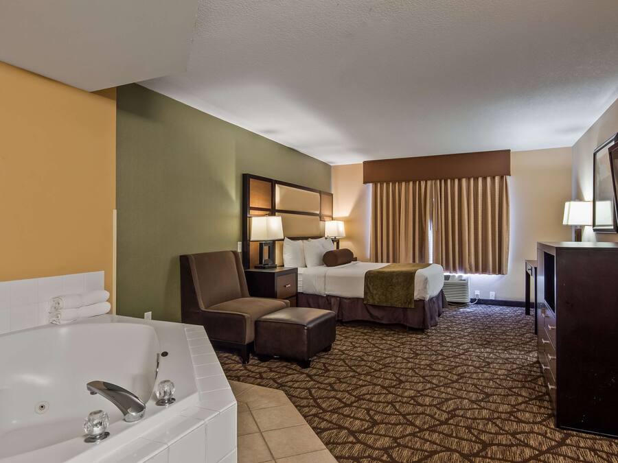Ein Hotelzimmer mit einer großen Badewanne, einem Bett mit zwei Kissen, einem Sessel, einem Schreibtisch, einem Fernseher und einem Fenster mit Vorhängen.