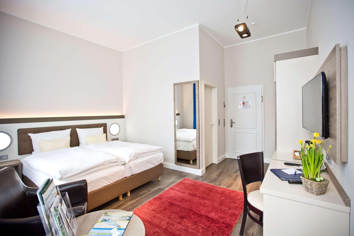 A hotel room with a bed, a desk, a chair, a red rug, and a mirror on the wall.