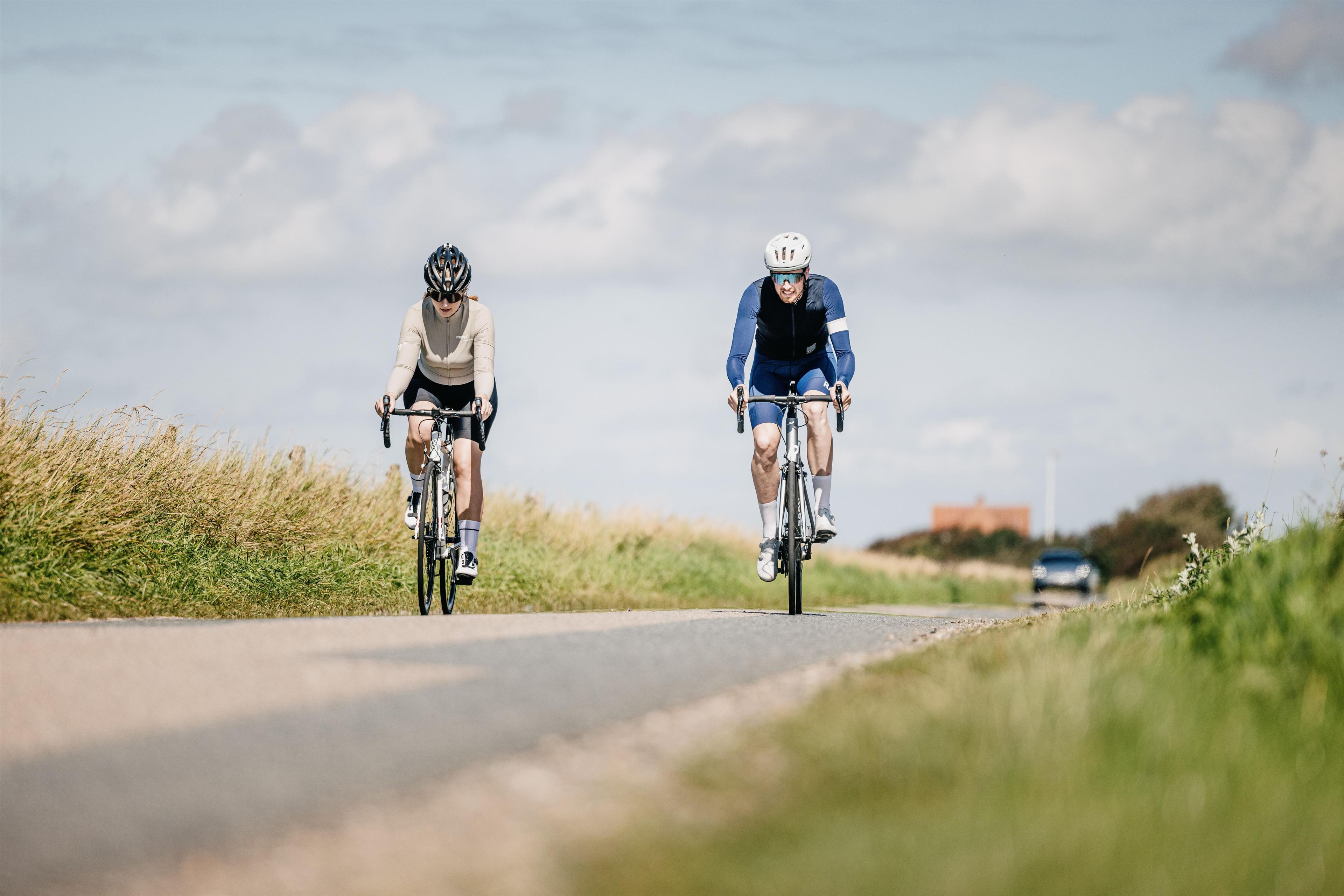 Deux cyclistes sur une piste asphaltée, de l’herbe de chaque côté, un ciel dégagé et un bâtiment visible au loin.