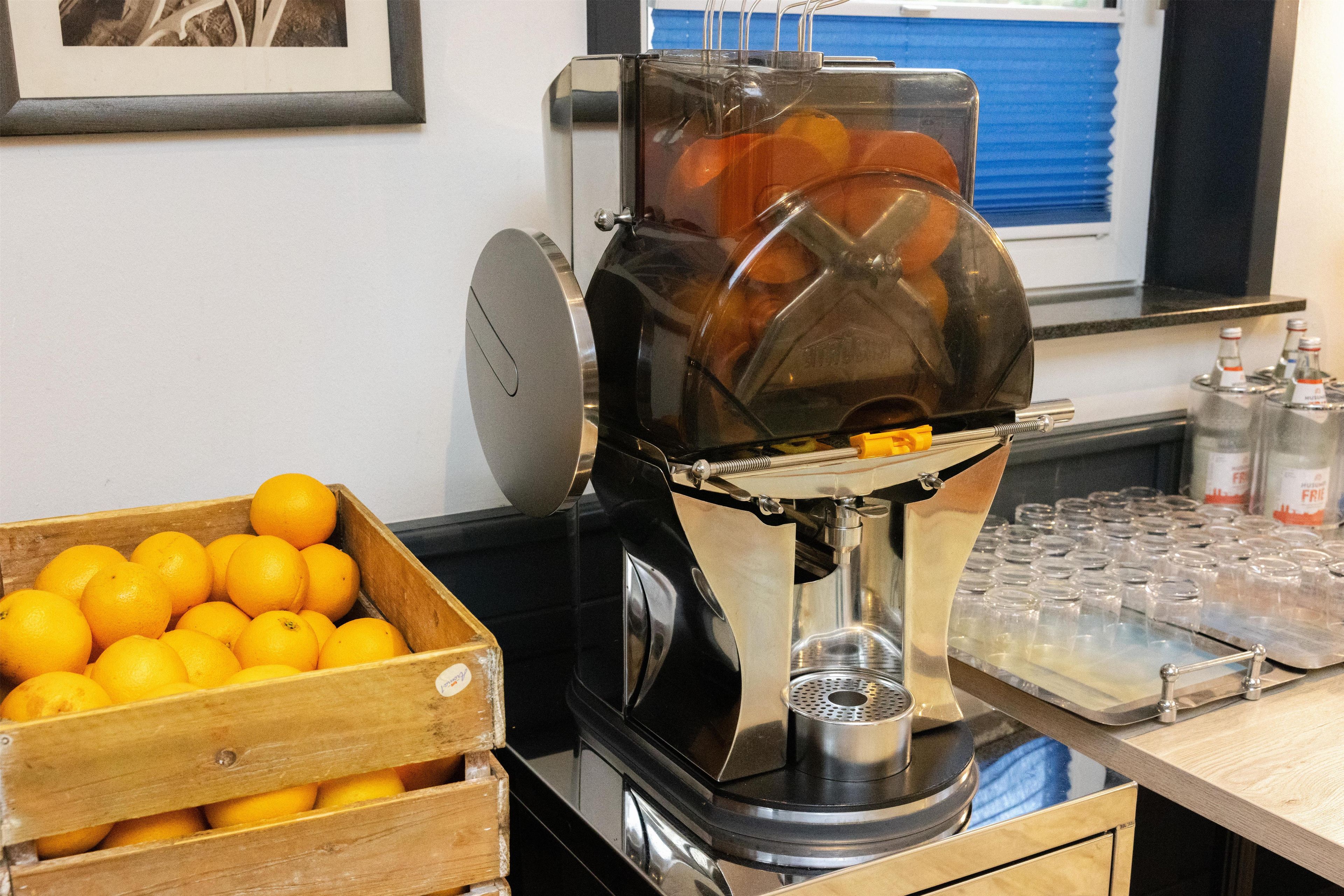 Una cafetera con un depósito de agua transparente, un cesto de madera lleno de naranjas y una bandeja de vasos vacíos.