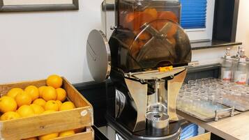 Una cafetera con un depósito de agua transparente, un cesto de madera lleno de naranjas y una bandeja de vasos vacíos.