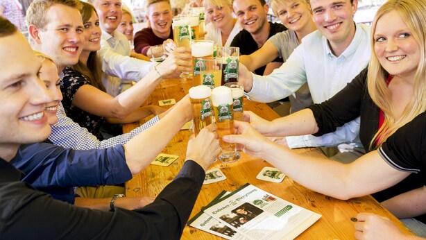 Un grupo de personas brindando con vasos de cerveza en una mesa de madera, con menús y un folleto a la vista.