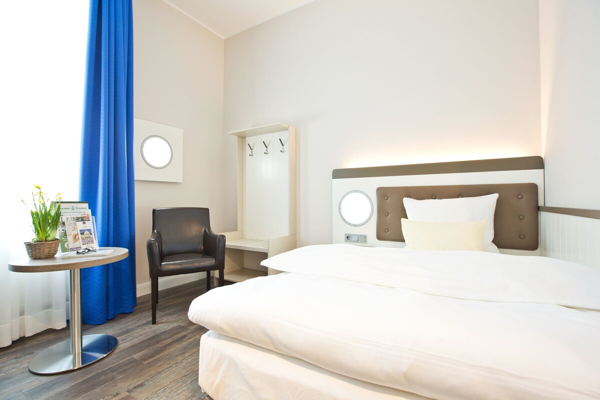 A hotel room with a bed, a chair, a small table with a plant, and a wall-mounted mirror.