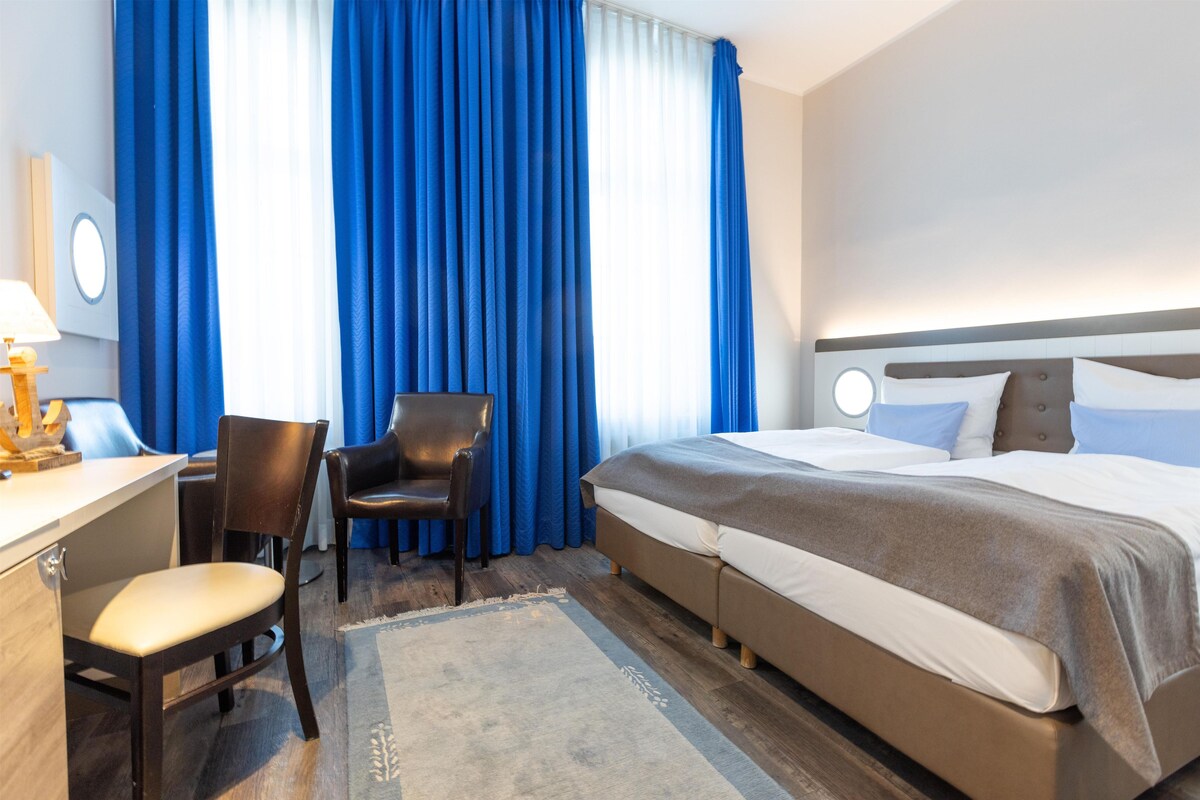 A hotel room with a bed, a desk with a chair, a lamp, and a window with blue curtains.