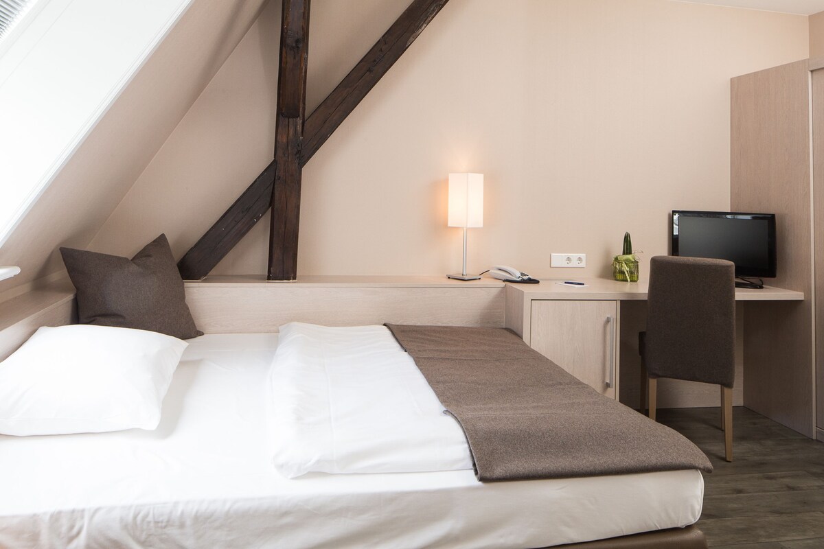 A modern hotel room with a bed, a desk, a chair, a TV, and a lamp.