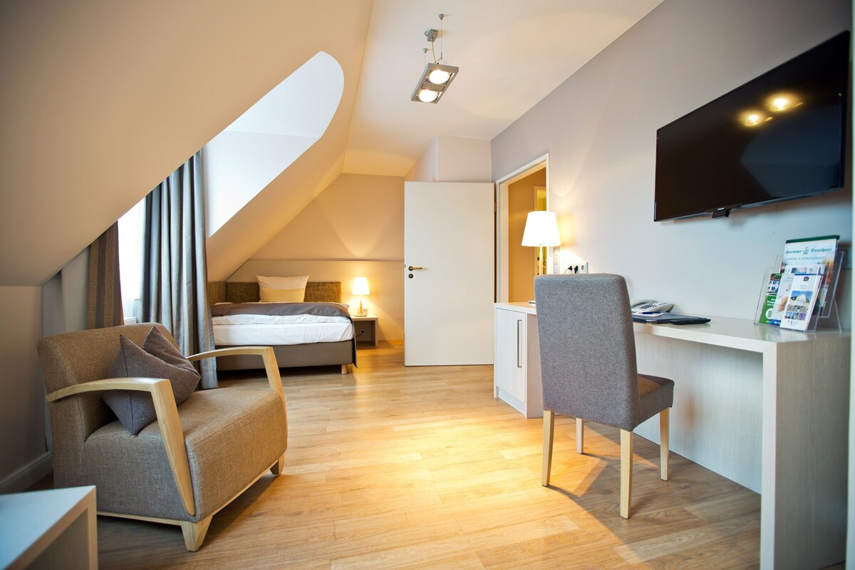 A modern hotel room with a bed, a desk with a chair, a TV, and a large window.