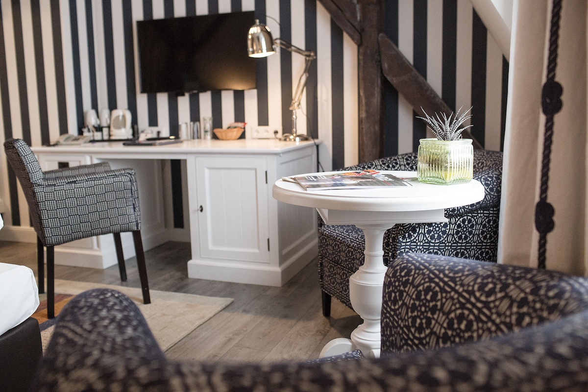 A room with a striped wall, a desk with a computer, a chair, a table with a magazine, and a decorative vase.