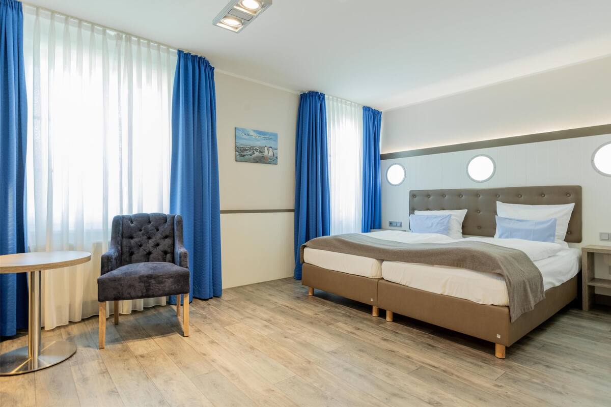 A hotel room with a bed, a chair, a small table, and a painting on the wall.