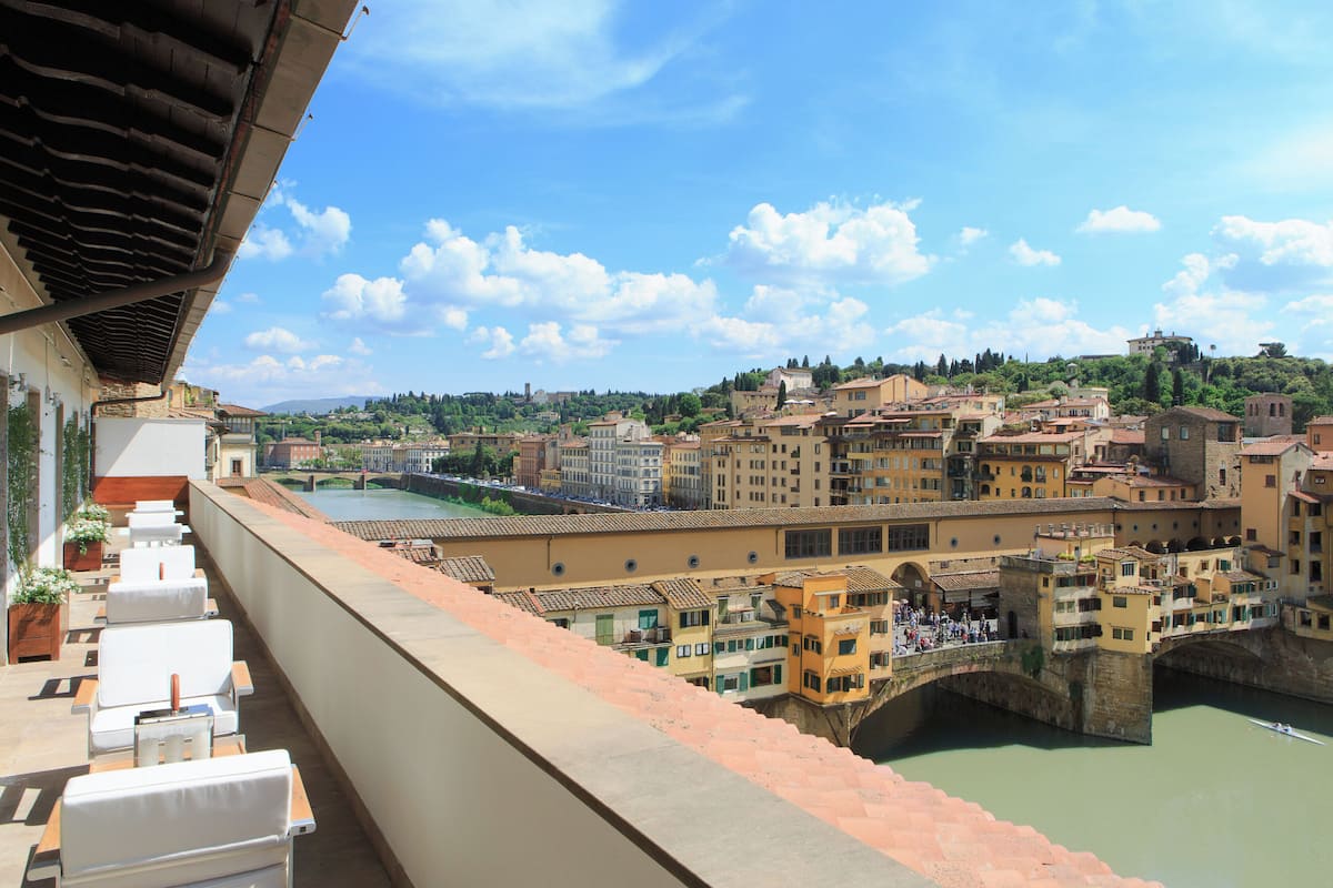 Un balcón con sillas blancas acolchadas con vistas a un pueblo histórico, un río y un puente.