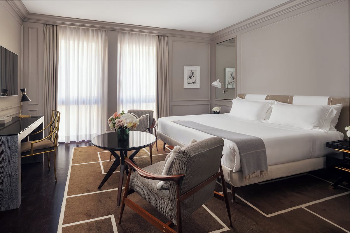Una habitación de hotel con una cama grande, un escritorio con una silla, una mesa redonda con un jarrón de flores y un ventanal con cortinas.