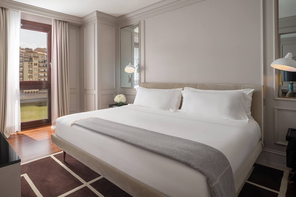 Un cuarto de hotel con una cama grande, una mesita de noche con lámpara, vista de edificios a través de una ventana y un espejo en la pared.
