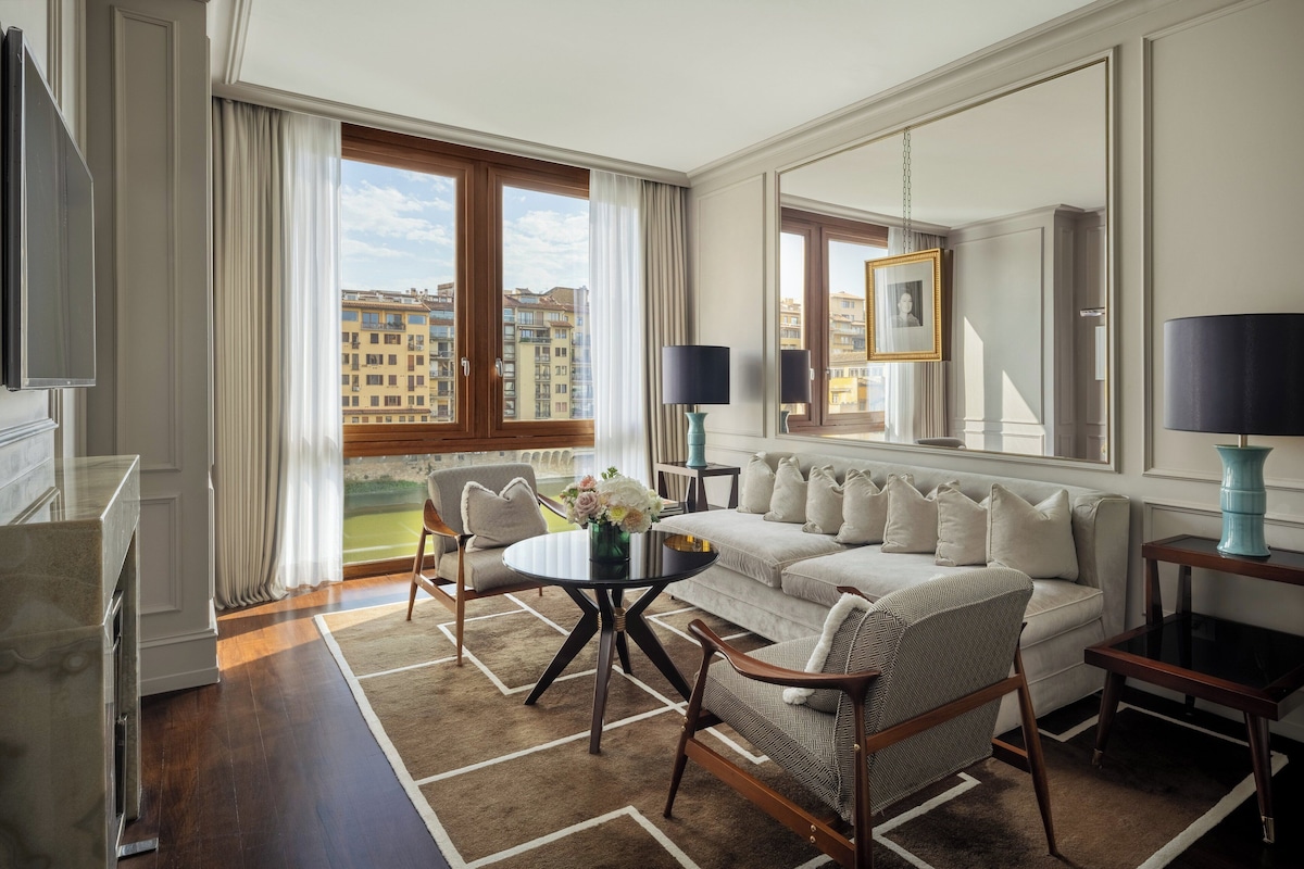 A modern hotel room with a sofa, chairs, a small table, and a large window offering a city view.