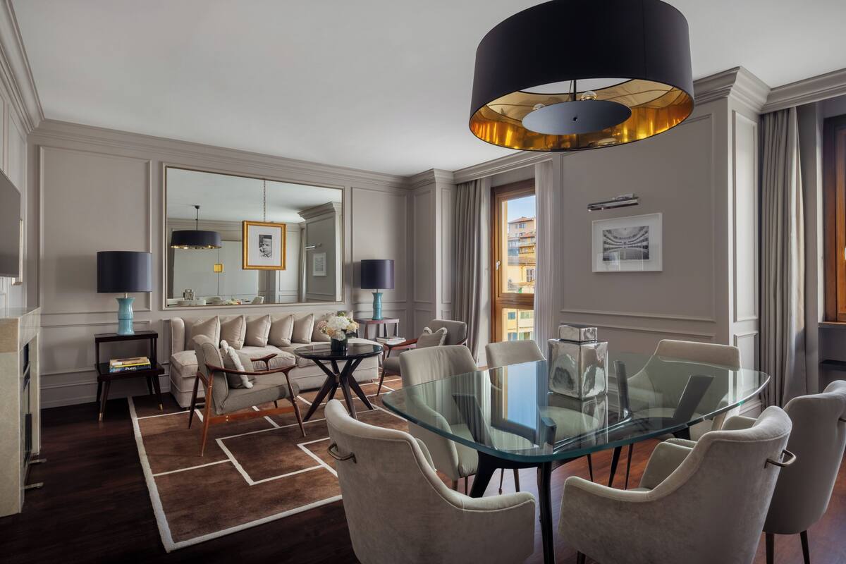 Una habitación de hotel moderna con una mesa de comedor de vidrio, asientos confortables, un espejo grande y vistas a la ciudad.