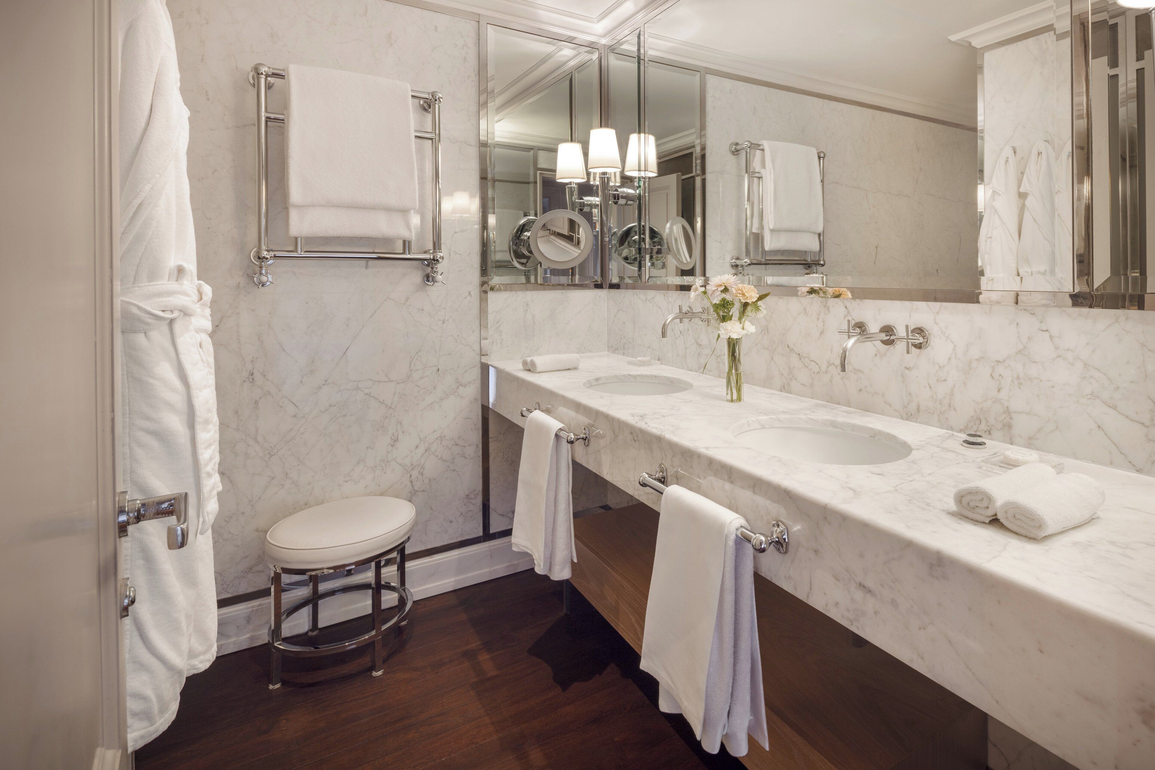 A modern bathroom with a marble countertop, two sinks, a vanity mirror, and a small stool.