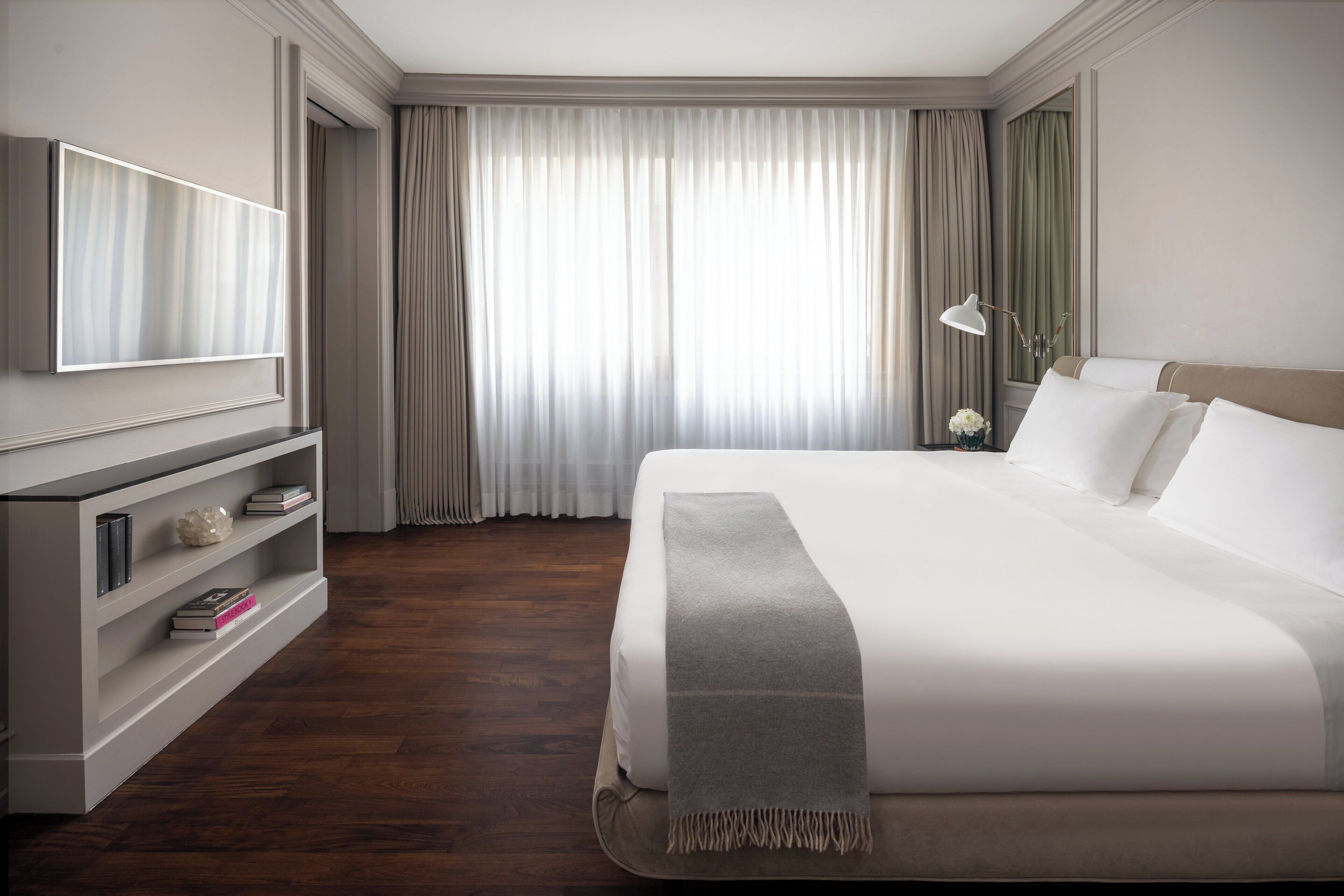 Een hotelkamer met een groot bed, een aan de muur bevestigde televisie, een boekenkast en een raam met gordijnen.