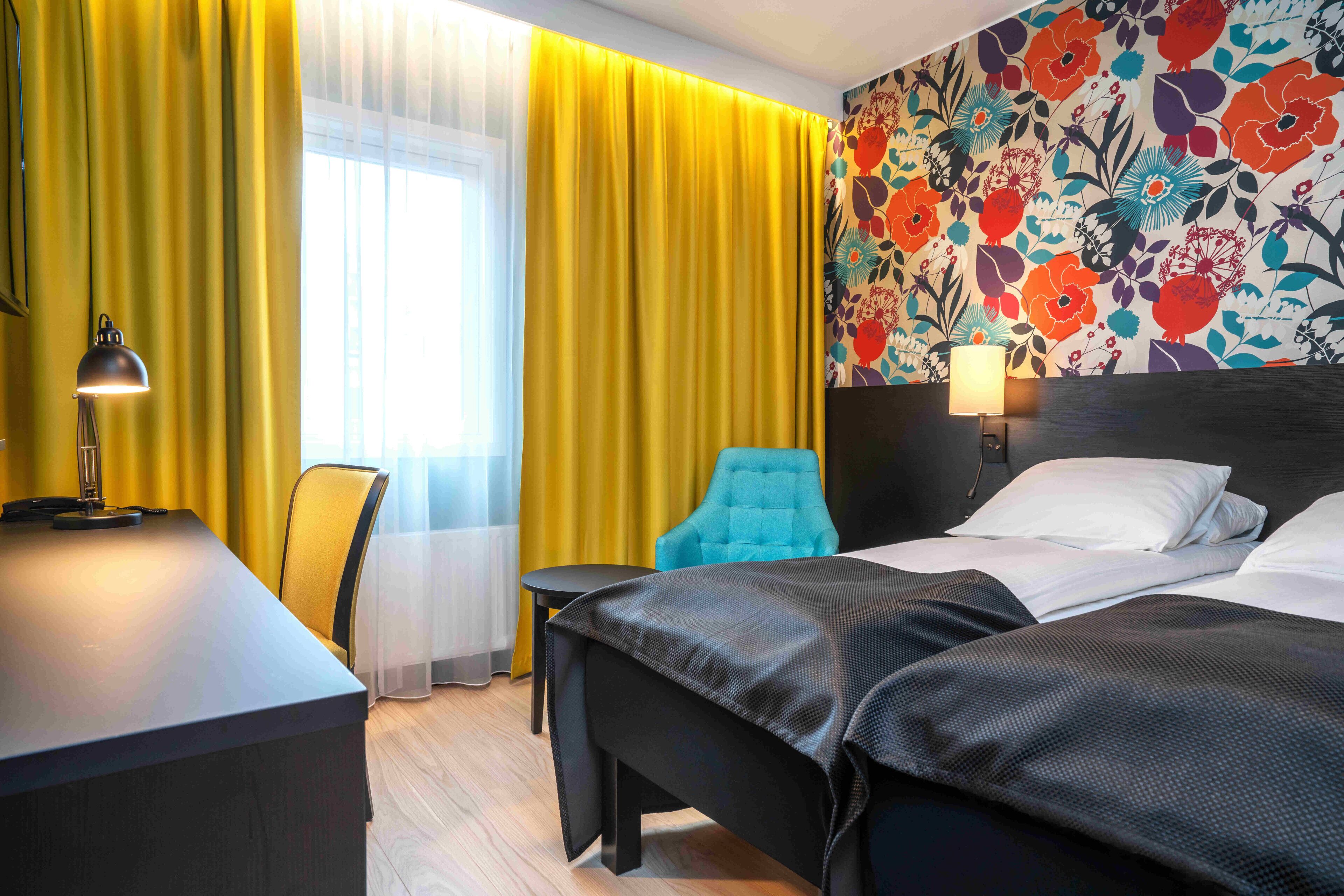 Ett hotellrum med en säng, ett skrivbord, en stol och ett fönster med gardiner.